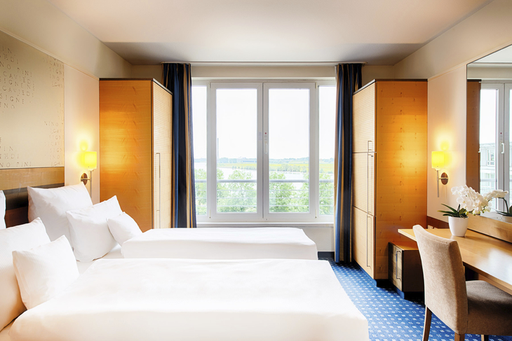 Helles Hotelzimmer mit zwei Betten, großem Fenster und Schreibtisch.