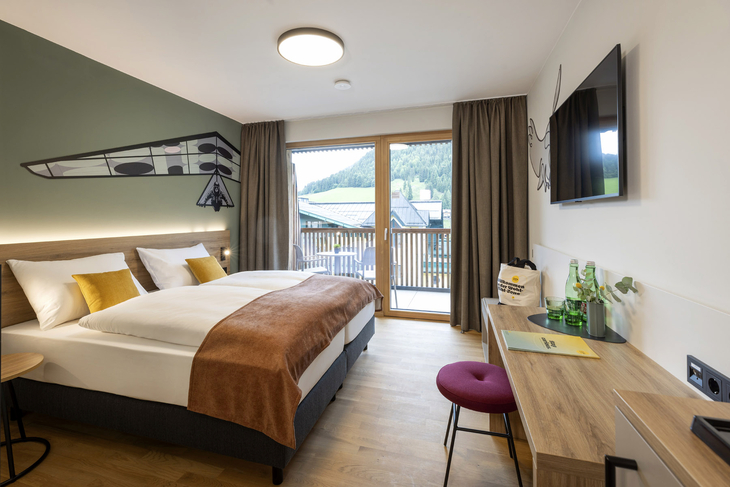 Hotelzimmer mit Doppelbett, Schreibtisch, TV und Balkonblick auf die Berge.