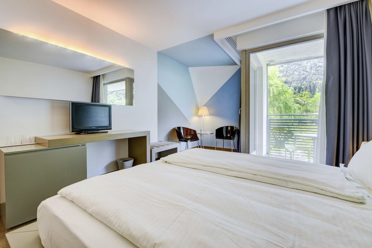 Helles Hotelzimmer mit Doppelbett, Fernseher und Balkon mit Ausblick.