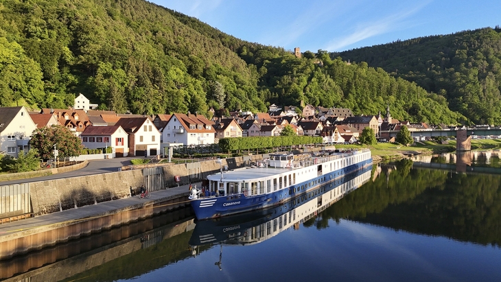 Flusskreuzfahrtschiff an einem sonnigen Tag vor einer bewaldeten Hügellandschaft und Dorf.
