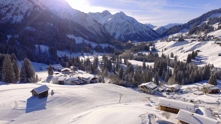 Verschneite Alpenlandschaft mit Hütten und Bergen im Hintergrund.