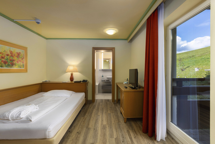 Einzelzimmer mit Bett, Lampe, TV, Balkon und Blick ins Grüne