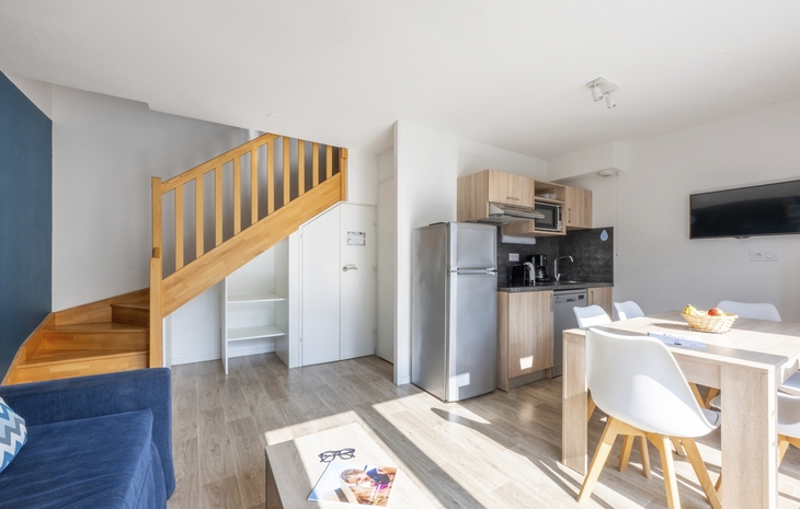 Helles Wohnzimmer mit Küche, Esstisch, Stühlen und Holztreppe.